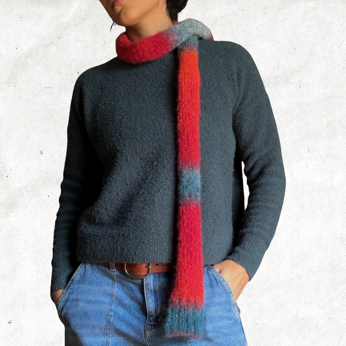 Front of a woman wearing a green sweater and jeans with a knitted skinny scarf that is made from the self striping red/orange/blue-grey yarn.