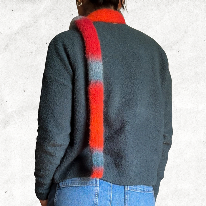 Back of a woman wearing a green sweater and jeans with a knitted skinny scarf that is made from the self striping red/orange/blue-grey yarn.